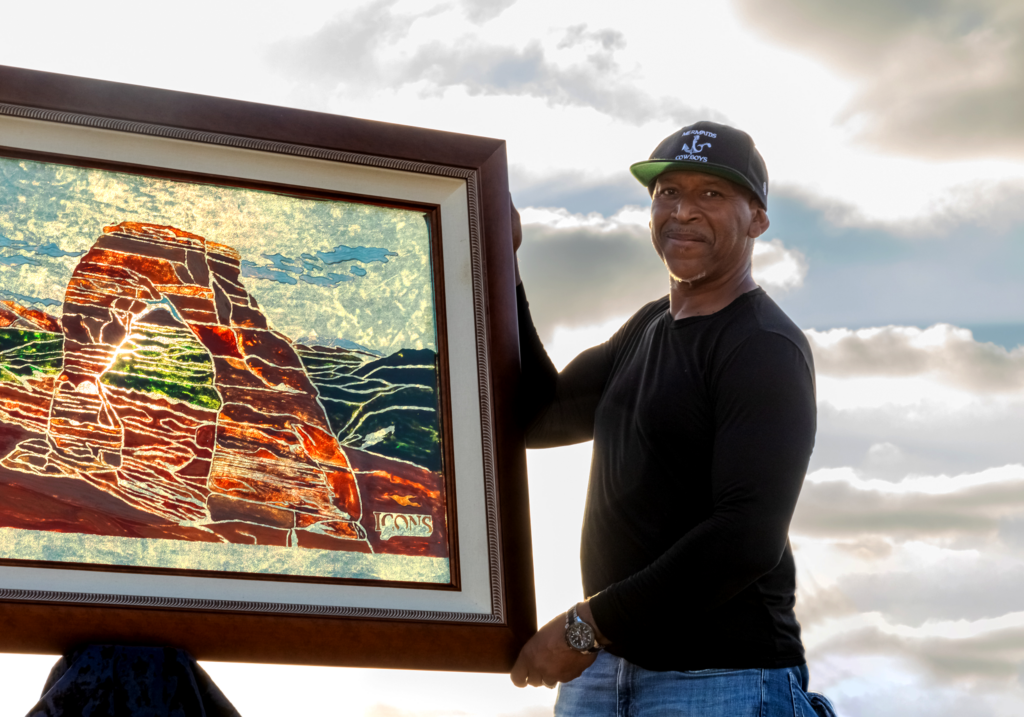 Greg “The Glassman” Holder holding a large framed artwork depicting a colorful landscape, standing outdoors under a cloudy sky - Icons by Holder | Sandblasted Glass Art