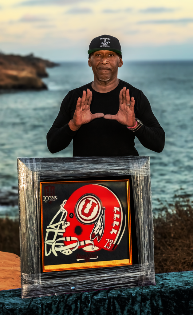A man, identified as Greg “The Glassman” Holder, stands outdoors near the ocean holding a framed glass artwork of a red football helmet with a white "U" logo - Icons by Holder | Sandblasted Glass Art