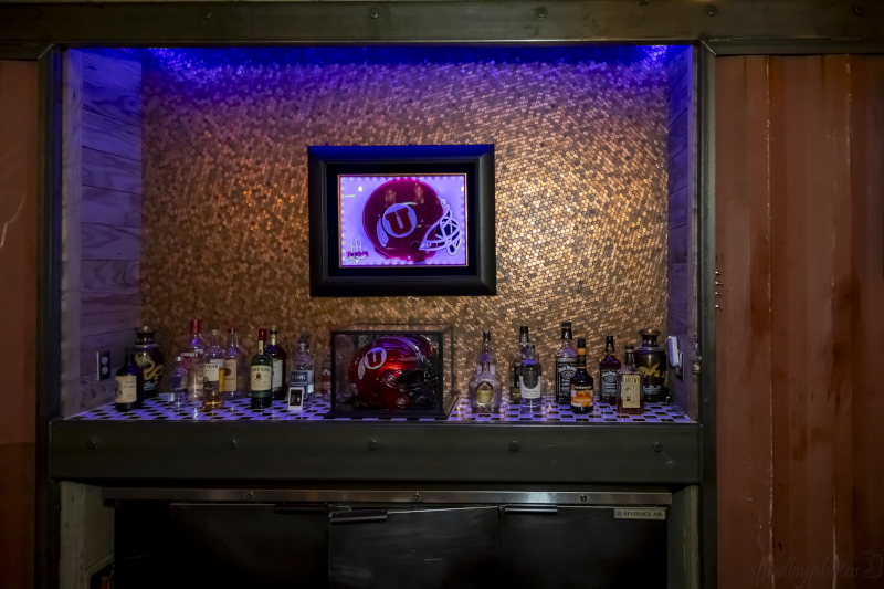 A display area features a framed artwork of a red football helmet with a white "U" logo, surrounded by bottles of alcohol on a countertop, under dim lighting with blue and amber accents - Icons by Holder | Sandblasted Glass Art